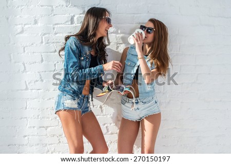 two pretty girls wearing sunglasses in summer jeanswear street urban casual style talking, laughing having fun on the background of brick wall