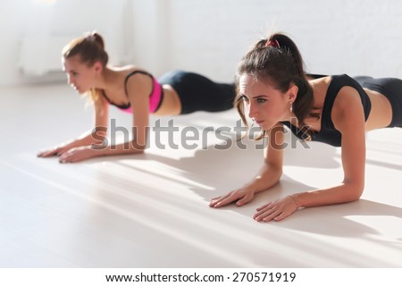 Two sporty fit and slim girls doing planking exercise indoors together with a natural light in sport studio hall or yoga class.