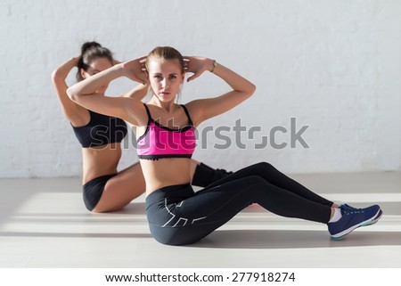 Women doing abdominal crunches stretching exercise hands behind heads at yoga class in fitness studio looking at camera.