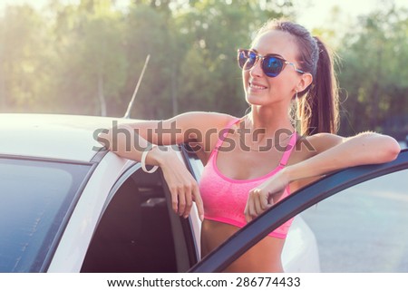Athlete sporty fit young woman in sports bra wearing sunglasses standing leaning on car with door open looking at camera