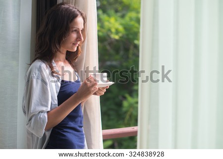 Woman in wide, long shirt is drinking tea or coffe and looking through the window.  Young lady is meeting sunrise