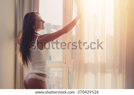 Lovely woman is awoke and standing before window. Girl opening curtains meeting sunrise