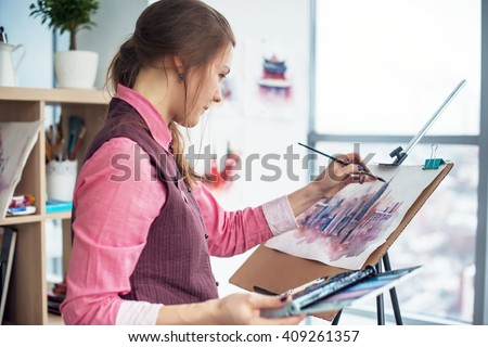 Side view portrait of a young woman painter drawing cityscape with watercolor palette on white paper using easel.
