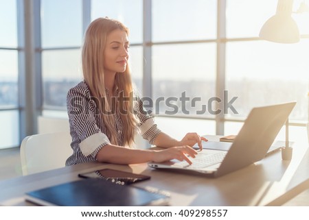 Young beautiful female copywriter typing texts and blogs in spacious light office, her workplace, using pc keyboard. Busy woman working.