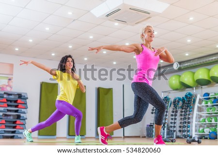 Two smiling female fitness models working out in gym or studio, doing cardio exercise, dancing zumba.