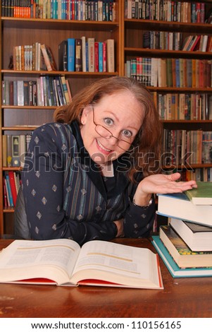 vertical orientation of a middle-aged woman in business attire with shallow depth of field, smiling and happy at a desk of books in a library / The Lifelong Learner