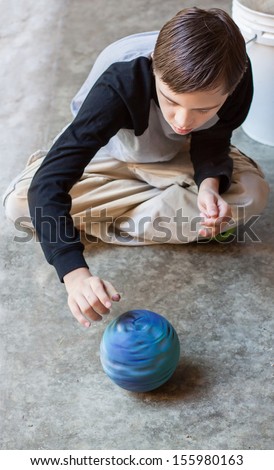 Vertical Orientation Of Boy With Autism And Down\'S Syndrome Sitting Cross Legged On A Cement Floor Spinning A Bright Blue Ball / Boy With Autism Spins A Ball