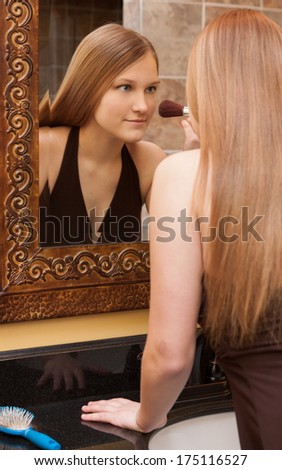 vertical orientation of a lovely teenage girl with long hair applying makeup with a brush as she looks into the mirror / Brushing up on Beauty