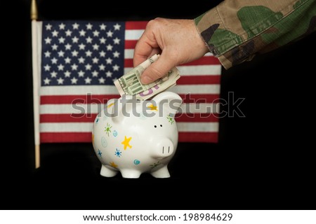 horizontal orientation close up of a woman's arm and hand in camouflage uniform putting cash in a piggy bank, with the U.S. flag in the background on isolated black / Saving while Serving - Horizontal