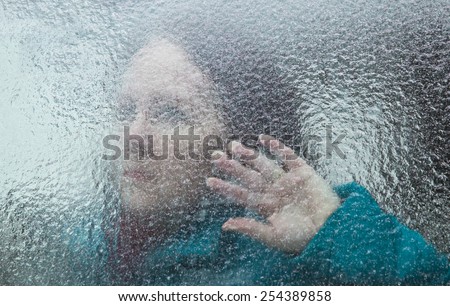 horizontal orientation of an attractive woman looking out through a window covered with thick ice, with her hand at the window / Signs of Alzheimers Disease