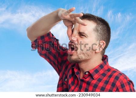 Young man with squares shirt over clouds background. Grabbing his nose with his hand