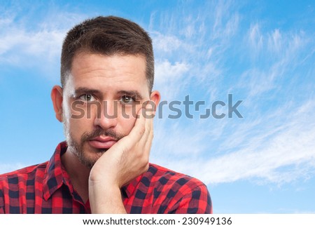 Young man with squares shirt over clouds background. Looking sad