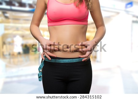 Young woman belly close up with a measurement scale. She is grabbing her fat. Over shopping center background