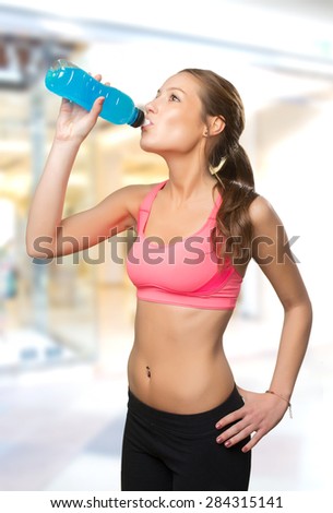 Young woman wearing gym clothes. She is drinking from a blue bottle. Over shopping center background