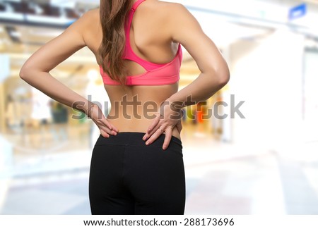 Young woman wearing a gym clothes. She has a back ache. Over shopping center background