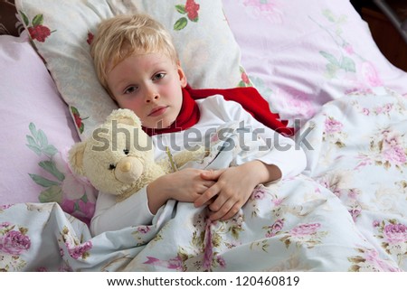 The little boy is sick. He lies in bed. Red scarf is on his neck. Teddy Bear is in his hand.