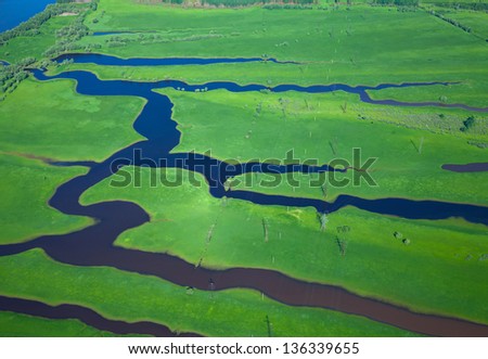 It is aerial view over the small river which is on the green meadow.