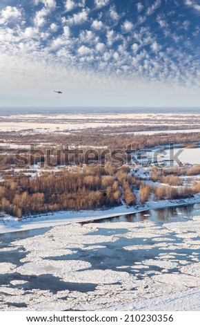 Helicopter is flying over snow covered plain beside the large river during spring break up. Helicopter MI-8 with external load. Aerial view.