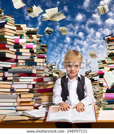 Little boy is reading interesting book. High stacks of books are on the table near him.