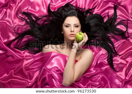 Beautiful girl with green apple and long black hairs lay on magenta fabric