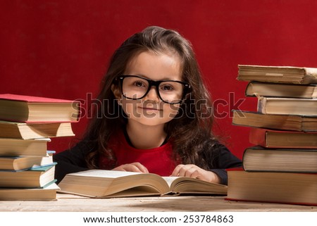 Cute little girl is reading a book while wearing glasses, isolated over red
