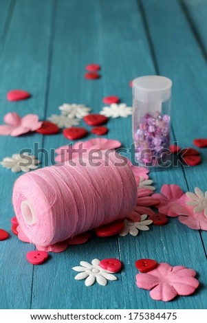 A reel of pink cotton thread with sewing accessories of buttons, beads and motifs, flowers and hearts, could be used for baby or valentine scrap booking,  on blue wooden boards