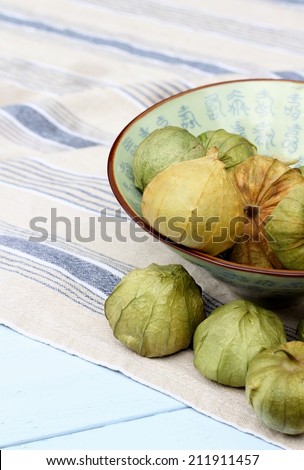 Ceramic bowl of green Tomatillos in their paper like outer skin, laying on blue striped cotton cloth towel, napkin