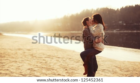 Young beautiful couple in love staying and kissing on the beach on sunset. Soft sunny colors.