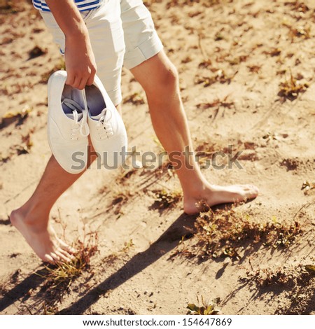 Young Man Walking On The Beach In Summer Vacation Carrying White Shoes In Hands