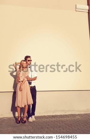 Young fashion elegant stylish couple posing on streets of european city in summer evening weather. Sensual blonde vogue girl with handsome hipster man posing near the big creamy wall.