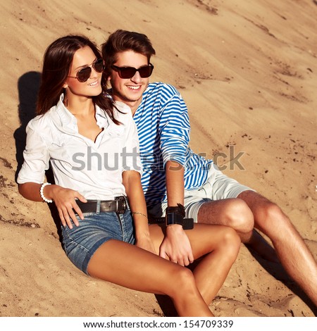 Pretty young fashion couple sitting on the beach having fun and looking at the sea in summer