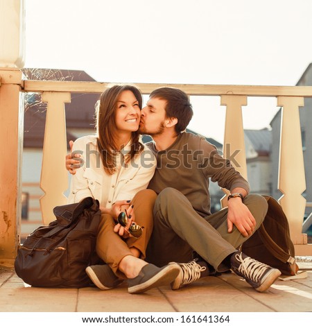 Young Beautiful Couple In Love Sitting And Posing Outdoor In Summer City While Traveling. Young Woman Smiling With Her Handsome Man