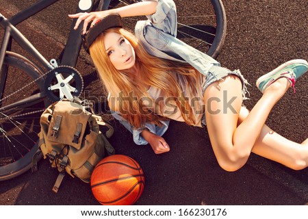 Outdoor summer fashion sensual portrait of beautiful long haired girl lying on the ground with bicycle and basketball ball sport swag style