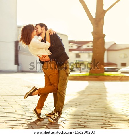 Pretty summer sunny outdoor portrait of young stylish couple while kissing on the street.