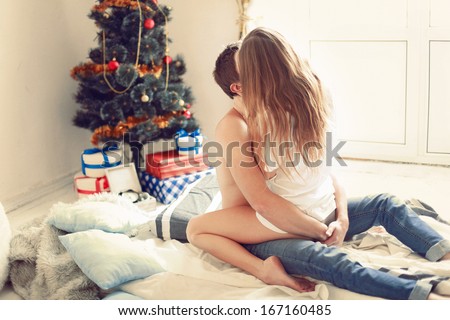 Young sweet pretty couple in love kissing in christmas eve near tree with gifts at home in december.