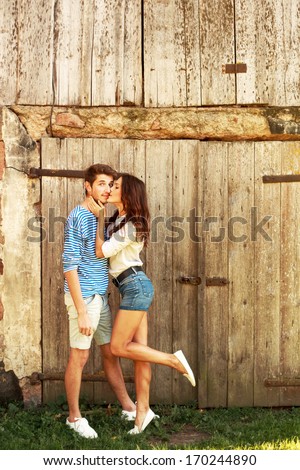 Young beautiful couple in love posing in spring near wooden door having fun and laughing. Girl kiss surprised man vintage style