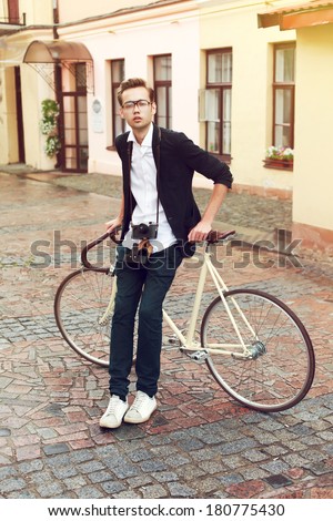 Outdoor portrait of young fashion hipster man posing on the street in old european city with bike and vintage camera in summer evening