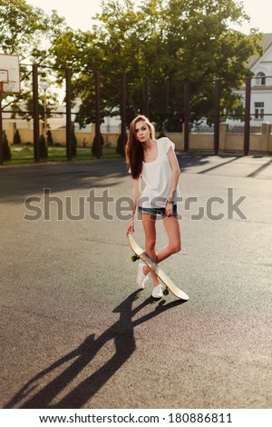 Young pretty fashion brunette girl posing on sport backyard of school in summer holiday with skateboard in hands. swag hipster style