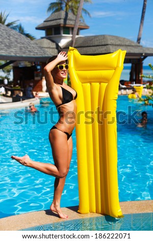 Summer outdoor portrait of pretty sporty young girl posing outdoor near pool with blue water and having fun with yellow mattress