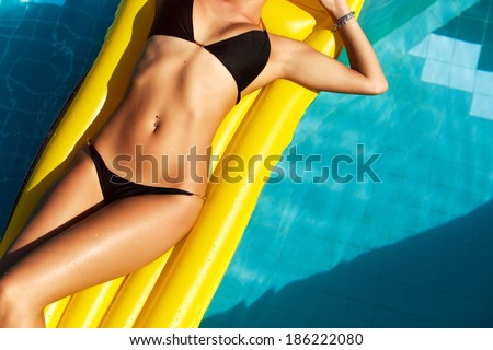 Young pretty woman with perfect tanned body lying on yellow air mattress in the pool in summer and having fun
