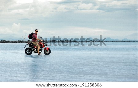 Young man traveling on motorcycle over the world standing on the sea behind blue water and sky and mountains feeling freedom on abandoned island with backpack