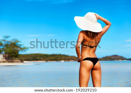 Outdoor summer portrait of pretty young tanned sexy hot woman standing back in bikini and sun hat on the beach and blue water and having fun on vacation