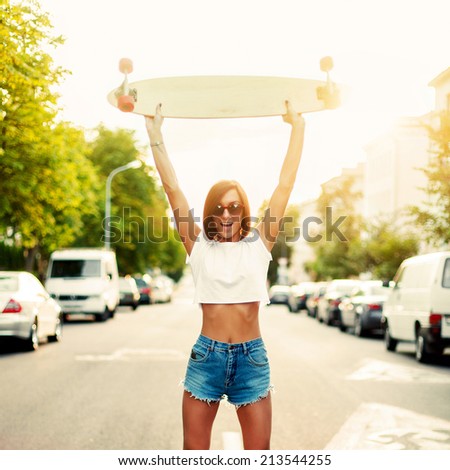 Pretty young happy girl posing with longboard hands up in the air in urban style evening have fun smiling dressed in jeans sort shorts