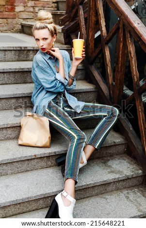 Outdoor fashion portrait of pretty young blonde sensual woman with drink glass and some food in paper pack have dinner on the street sitting on stairs