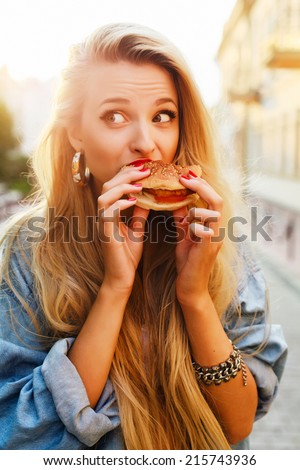 Young pretty funny fashion sensual blond girl eats hamburger fast food sandwich on the street lifestyle outdoor photo