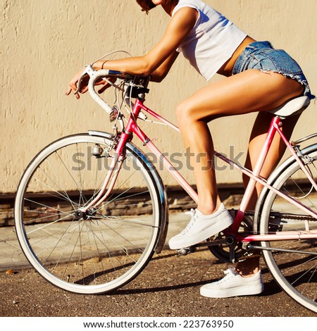 Outdoor portrait of pretty sexy young tanned sport style hot legs woman posing on pink bicycle on creamy wall background
