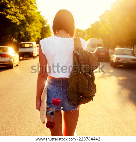 Pretty young sexy hot woman in short jeans shorts and hot body walking outdoor on the road in summer evening with backpack and longboard