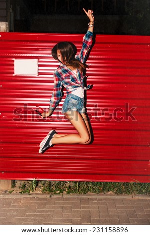 Outdoor night flash portrait of pretty sexy tanned young girl jumping going crazy and happy fun on the street on red wall background