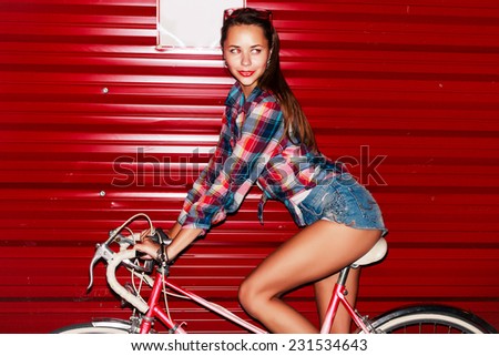 Pretty young sexy tanned sporty girl rides pink vintage bicycle in the night on red wall background