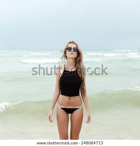 Sexy young blonde woman posing alone on the sea in summer dressed in black bikini and sunglasses walking on the beach behind blue waves of storm sea